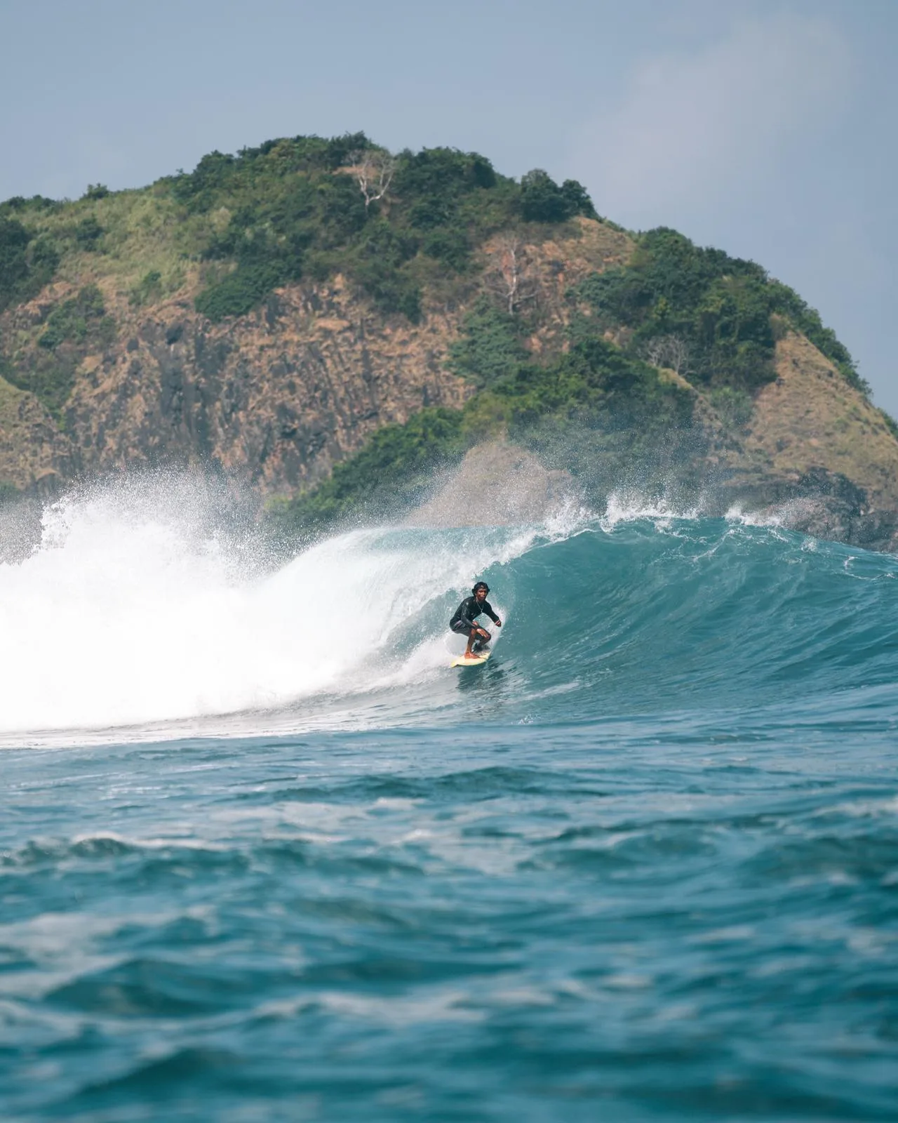 A surfer riding a turquoise wave beneath the hills of Lombok